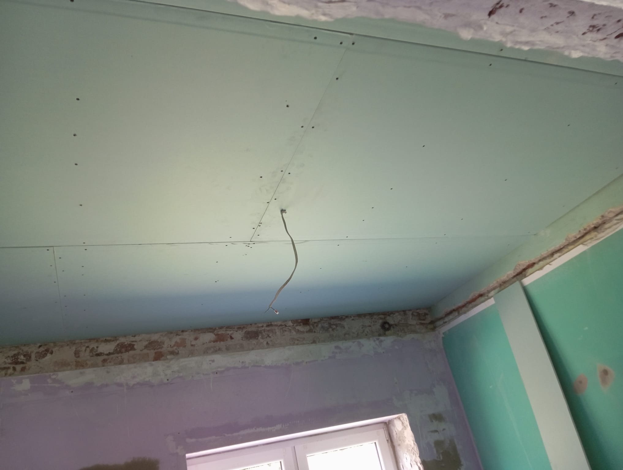 Montaż płyty G-K na suficie w trakcie remontu. Widoczne łączenia płyt, wystający kabel i surowy stan ścian. Ujęcie z dołu, podkreślające etap prac.