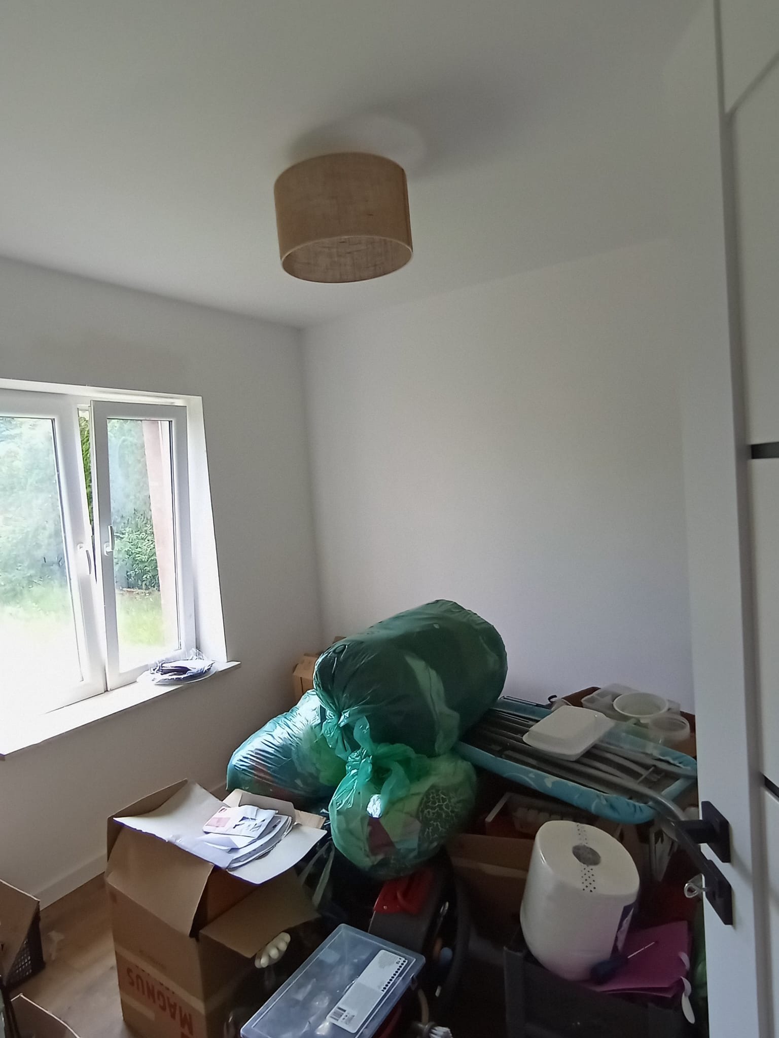 Pokój przed remontem: białe ściany, okno z widokiem na zieleń, pod sufitem lampa. Na podłodze kartony, worki ze śmieciami i inne przedmioty, tworzące bałagan.