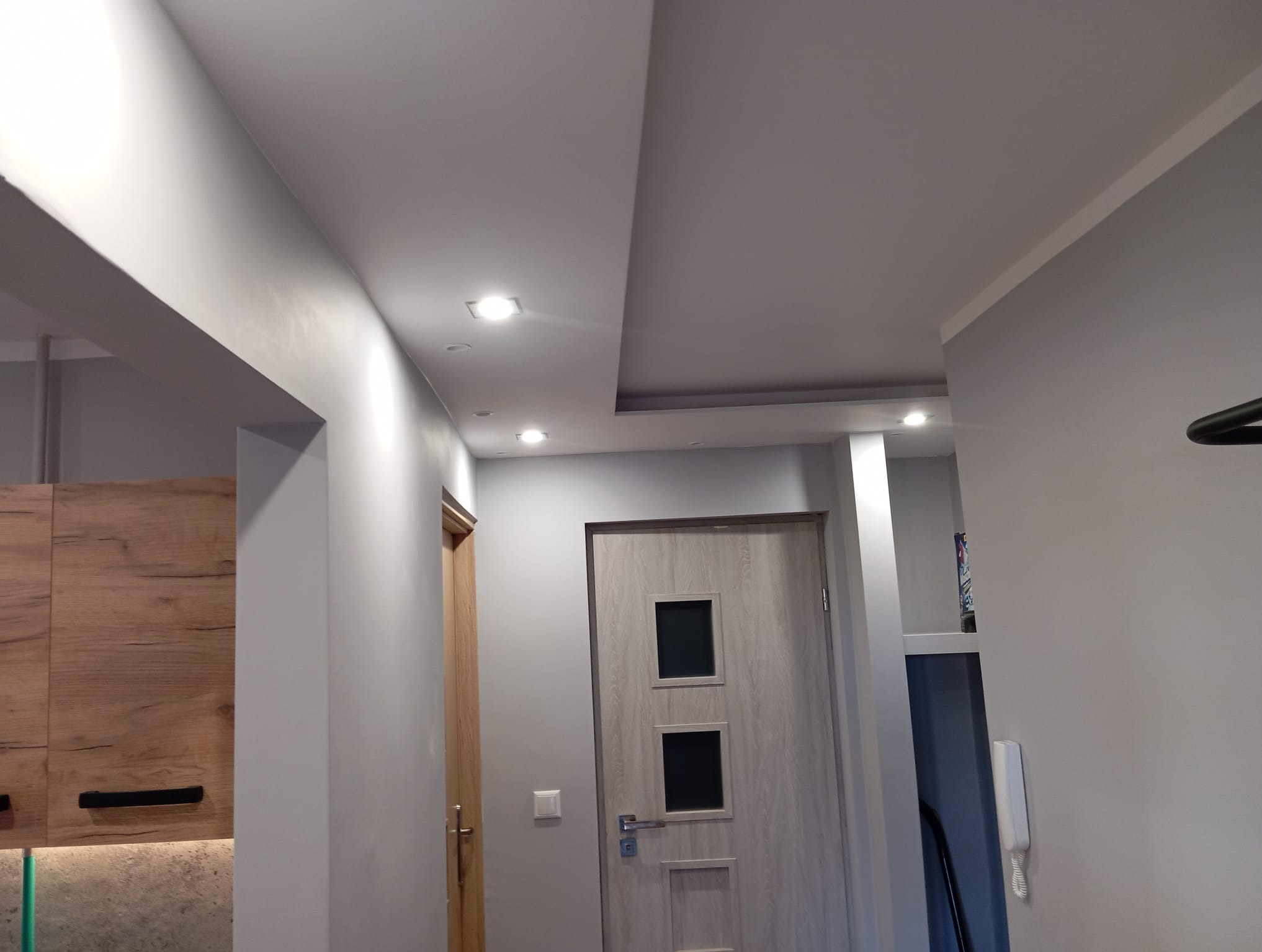 Nowoczesny korytarz po remoncie: szare ściany, podwieszany sufit z oświetleniem punktowym, drewniane drzwi i zabudowa meblowa. Minimalistyczny design.
