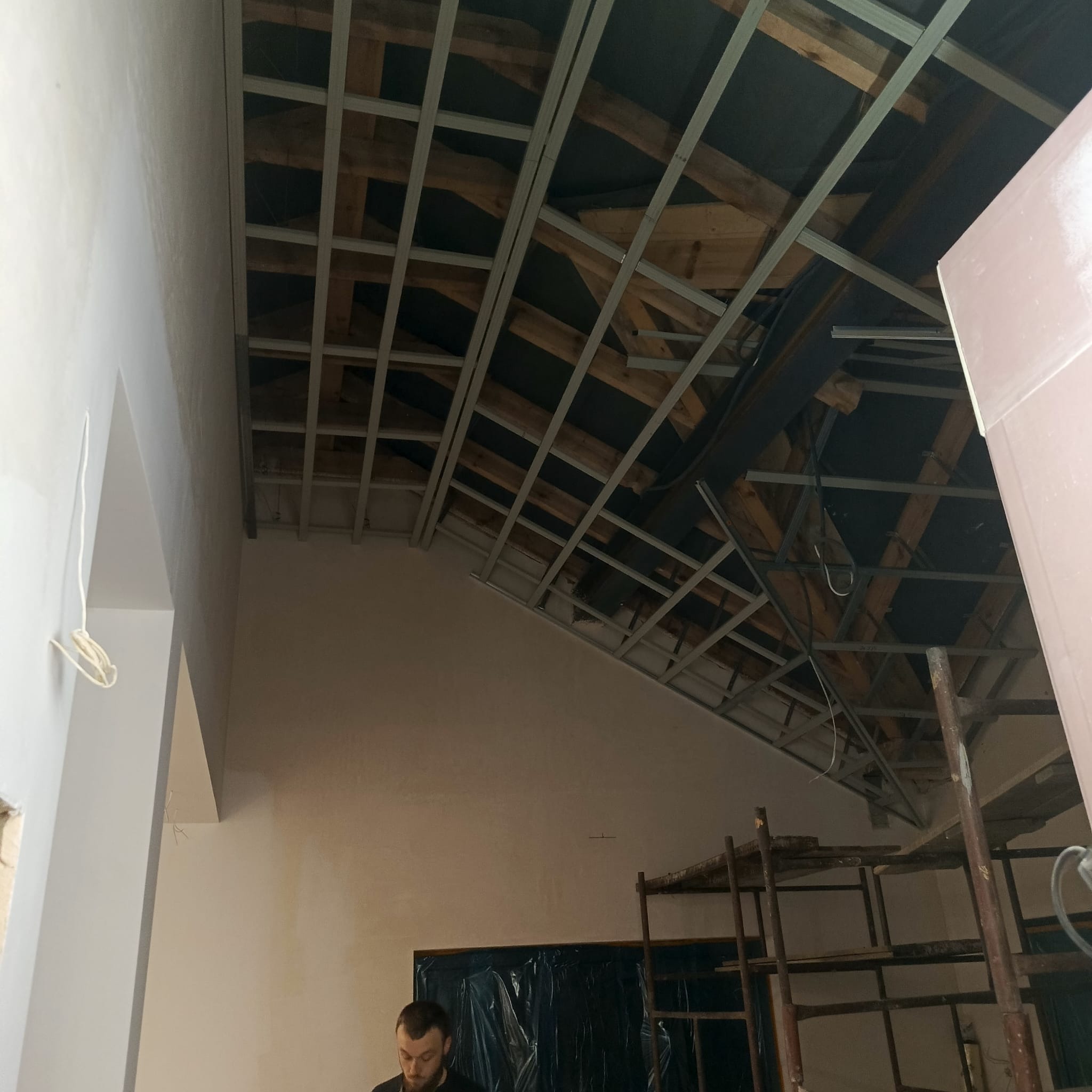 Skośny sufit z metalową konstrukcją pod płytę G-K, częściowo oszpachlowana ściana, rusztowanie i folia ochronna. Widoczny fragment osoby w dolnej części kadru.