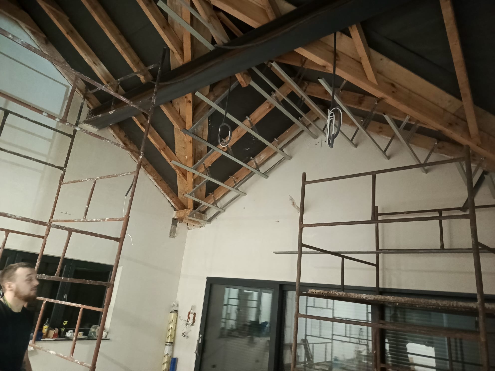 Skośny sufit z drewnianą konstrukcją i metalowym stelażem pod płyty. Widoczne rusztowania i fragment osoby. Prace wykończeniowe w jasnym pomieszczeniu.