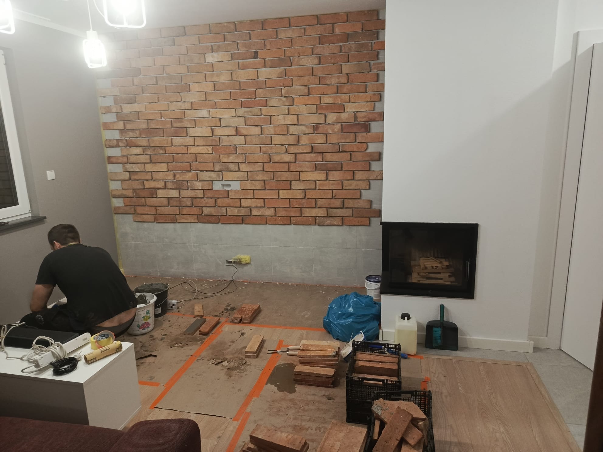 Wnętrze w trakcie prac: mur z cegły klinkierowej na ścianie, widoczny kominek, narzędzia i materiały budowlane, osoba w trakcie pracy. Ujęcie z perspektywy podłogi.