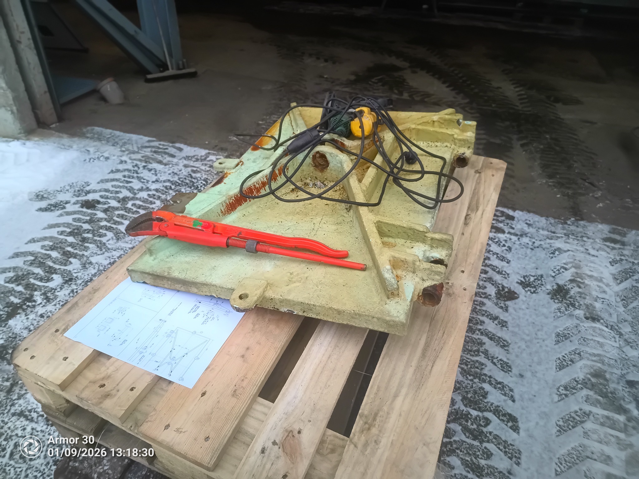 Żółta, metalowa konstrukcja na palecie z narzędziami i rysunkiem technicznym. W tle ślady opon na śniegu, sugerujące odśnieżanie dróg i placów.