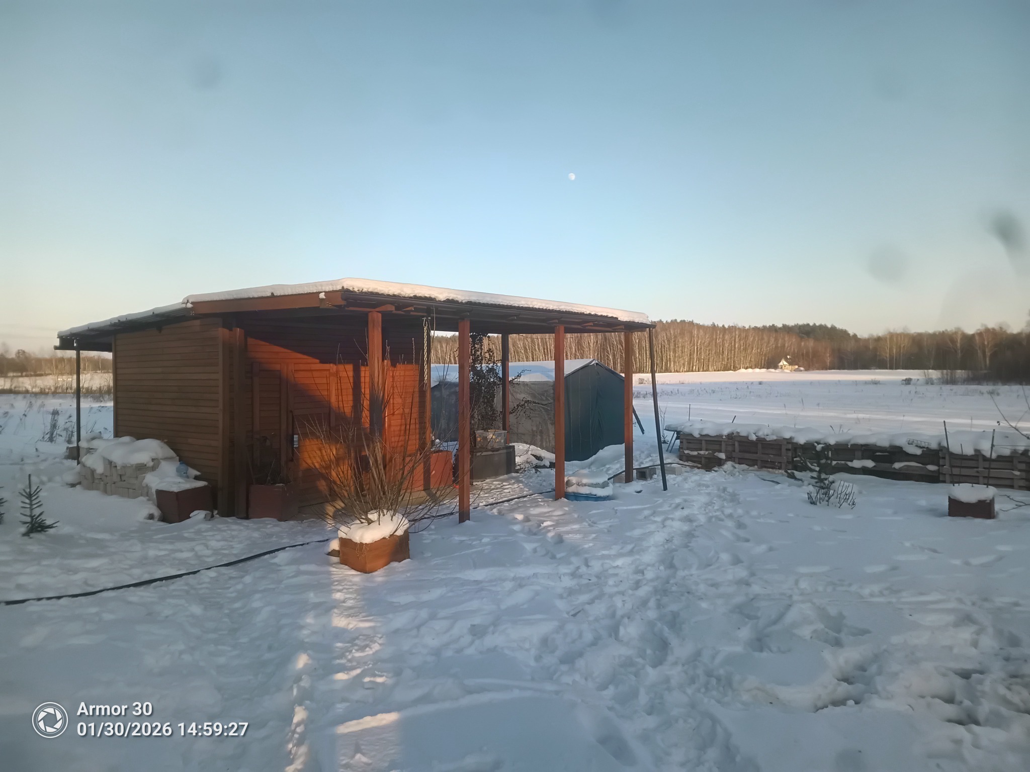 Zimowy krajobraz z drewnianą altaną i zasypanym śniegiem ogrodem. W tle widoczny las i błękitne niebo z księżycem. Ujęcie w słoneczny dzień.