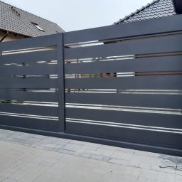 Hubert Złotogórski - Nowoczesna, przesuwna brama w kolorze grafitowym z poziomymi, ażurowymi przerwami, zamontowana na posesji z kostki brukowej i elementami domu w tle.