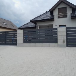 Hubert Złotogórski - Nowoczesne ogrodzenie posesji z szarego metalu i betonu architektonicznego, brama wjazdowa i furtka, na tle domu z ciemną dachówką i kostki brukowej.