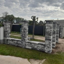 Hubert Złotogórski - Fragment ogrodzenia z szarej kostki łupanej, z widoczną czarną siatką w tle i pochmurnym niebem. Elementy ogrodzenia w surowym stylu, z wyraźną fakturą kamienia.