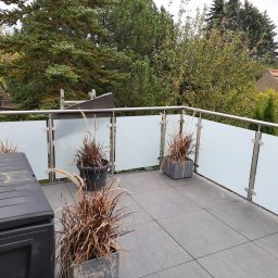 Hubert Złotogórski - Nowoczesna balustrada balkonowa ze stali nierdzewnej i matowego szkła, z donicami z ozdobnymi trawami na szarych płytkach. Widok na zieleń drzew i fragment dachu.