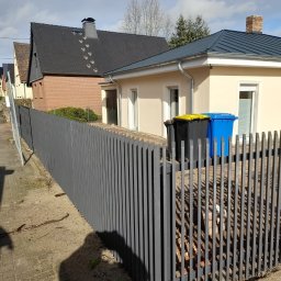 Hubert Złotogórski - Nowoczesne, szare ogrodzenie palisadowe z metalu, widoczne na tle domów jednorodzinnych i pojemników na śmieci. Ujęcie z poziomu chodnika.