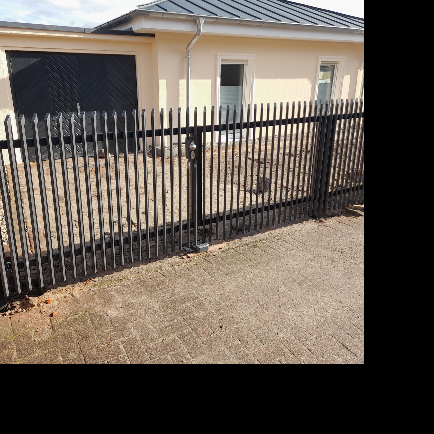 Nowoczesne, metalowe ogrodzenie palisadowe w kolorze grafitowym, z widoczną furtką i zamkiem, na tle beżowego budynku z ciemną bramą garażową.