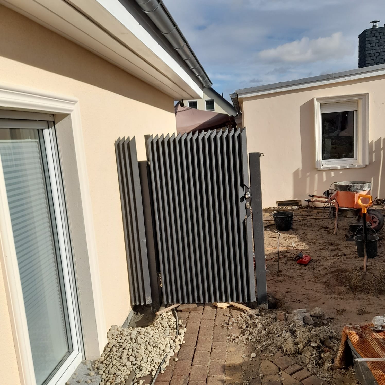 Nowoczesna, metalowa furtka z pionowymi szczeblami, zamontowana przy budynku z jasną elewacją. Widoczne narzędzia i materiały budowlane sugerują etap montażu.