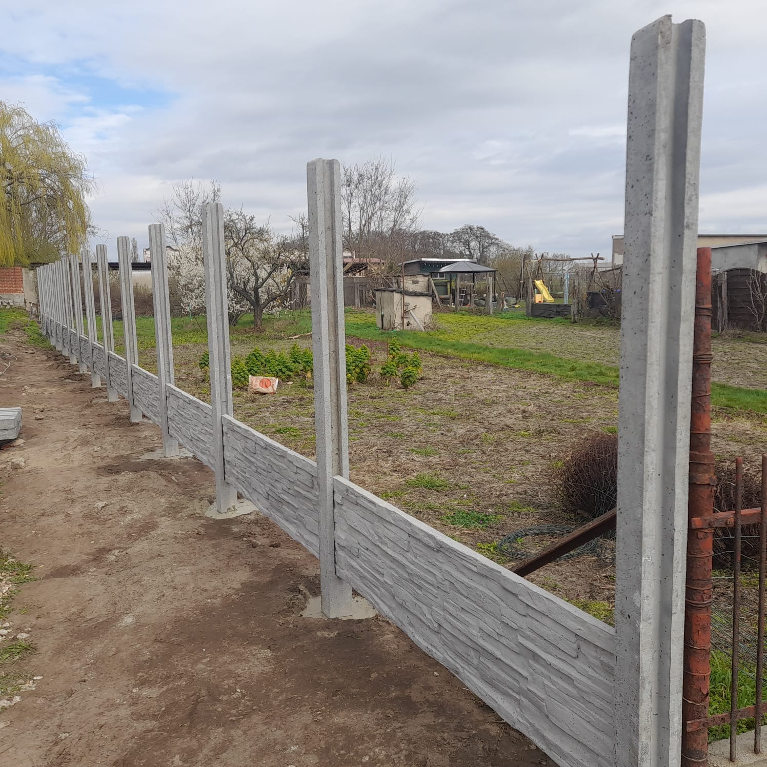 Montaż prefabrykowanego ogrodzenia betonowego z imitacją kamienia. Widoczne słupki i panele w trakcie instalacji, surowy krajobraz w tle.