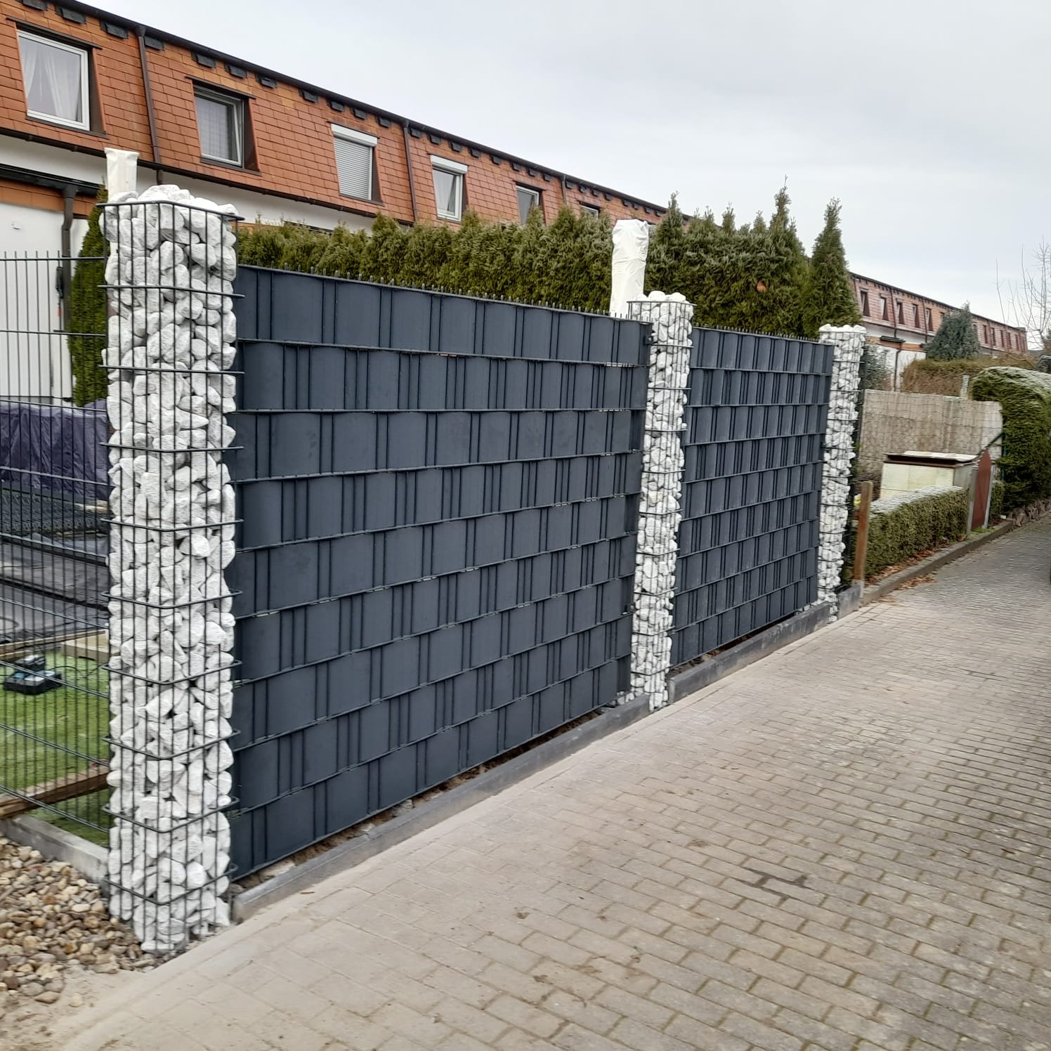 Nowoczesne ogrodzenie panelowe w kolorze grafitowym, z filarami wypełnionymi białymi kamieniami. Widok z poziomu chodnika, w tle budynek mieszkalny.