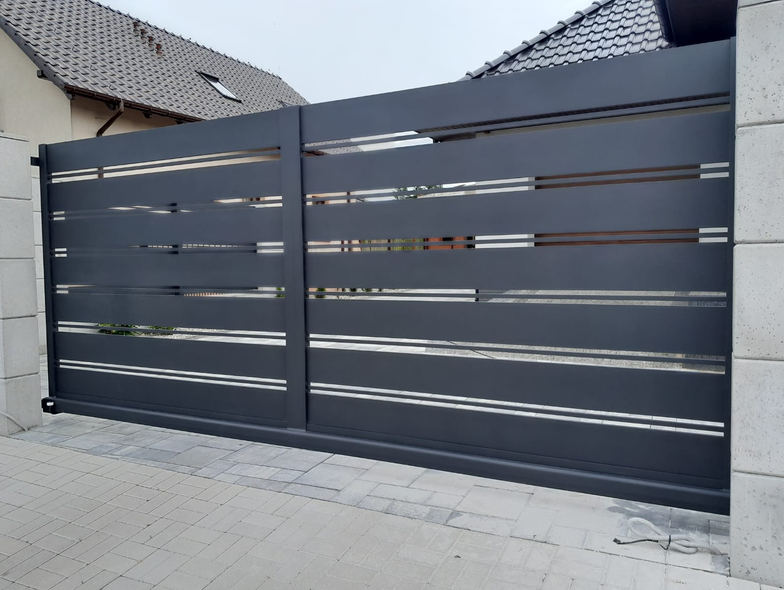 Nowoczesna, przesuwna brama w kolorze grafitowym z poziomymi, ażurowymi przerwami, zamontowana na posesji z kostki brukowej i elementami domu w tle.
