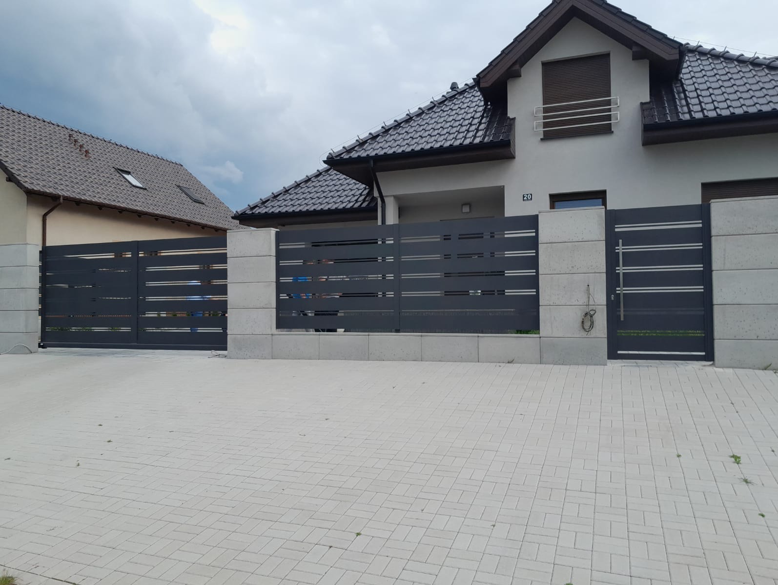 Nowoczesne ogrodzenie posesji z szarego metalu i betonu architektonicznego, brama wjazdowa i furtka, na tle domu z ciemną dachówką i kostki brukowej.