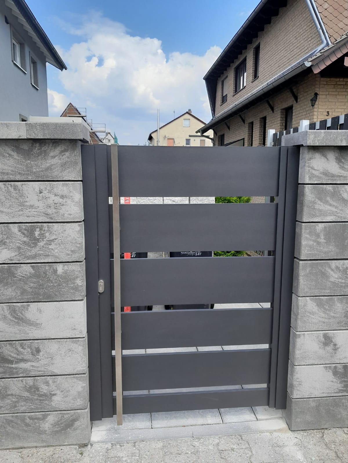 Nowoczesna, szara brama z poziomych paneli, osadzona w słupkach z szarej kostki. Widoczne detale zamka i metalowego wzmocnienia. Tło stanowią budynki mieszkalne i błękitne niebo.