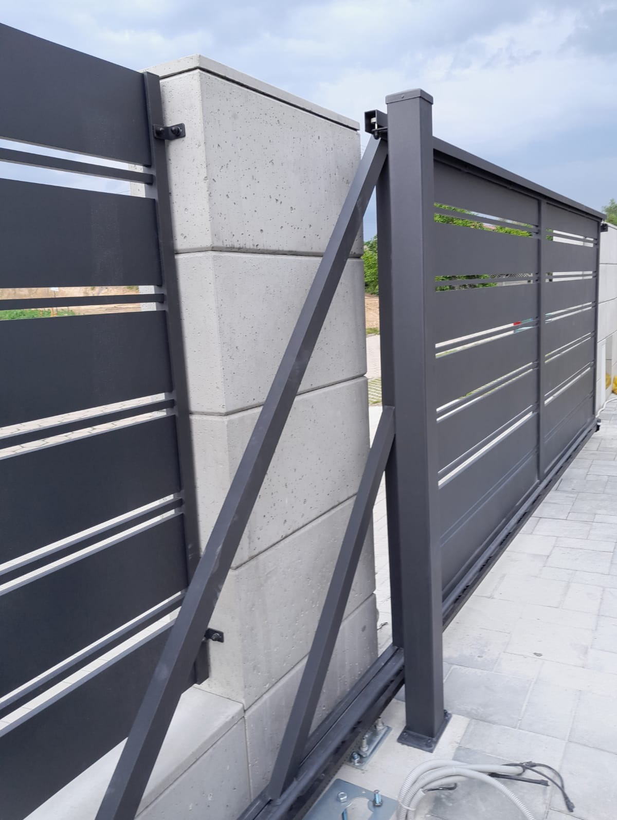 Nowoczesna, przesuwna brama w kolorze grafitowym z poziomymi szczelinami, zamontowana na betonowym słupku. Widoczne elementy mocujące i fragment kostki brukowej.