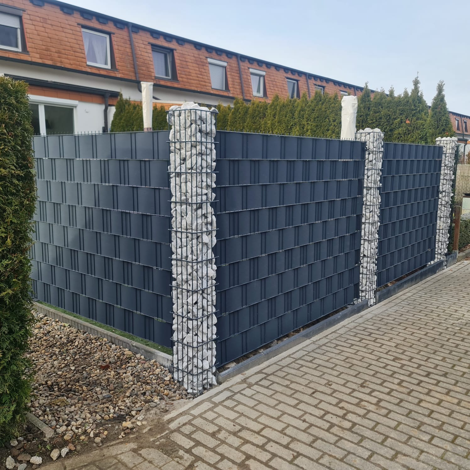 Nowoczesne ogrodzenie panelowe w kolorze grafitowym z gabionowymi słupkami wypełnionymi białymi kamieniami, oddzielające posesję od chodnika.