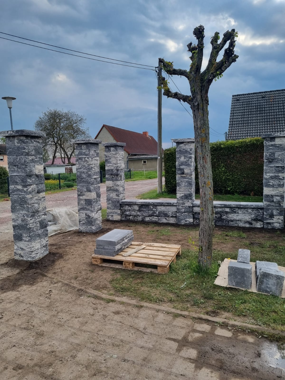 Wznoszenie murowanego ogrodzenia z szarej kostki. Widoczne słupki i fragment muru w trakcie budowy. Na pierwszym planie paleta z kostką i drzewo.