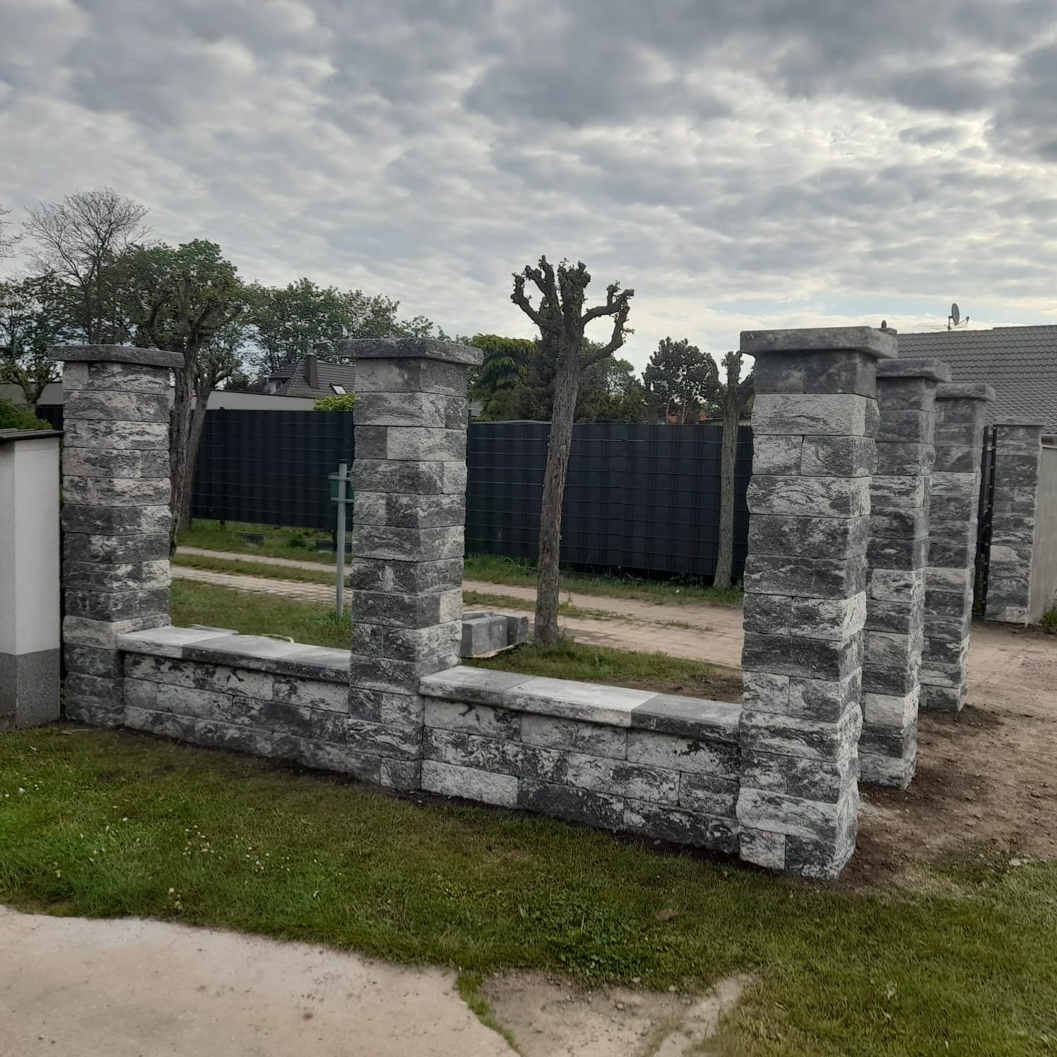 Fragment ogrodzenia z szarej kostki łupanej, z widoczną czarną siatką w tle i pochmurnym niebem. Elementy ogrodzenia w surowym stylu, z wyraźną fakturą kamienia.