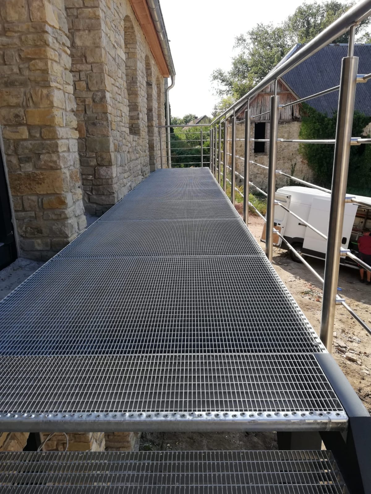 Metalowa kładka z kraty pomostowej i balustradą ze stali nierdzewnej, prowadząca wzdłuż kamiennej ściany budynku. Widok z dołu, na zewnątrz.