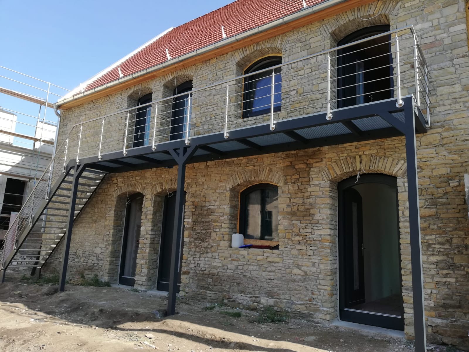 Kamienny budynek z metalową konstrukcją balkonu i schodami. Nowoczesne balustrady ze stali nierdzewnej kontrastują z rustykalnym stylem budynku. Widoczne detale architektoniczne.