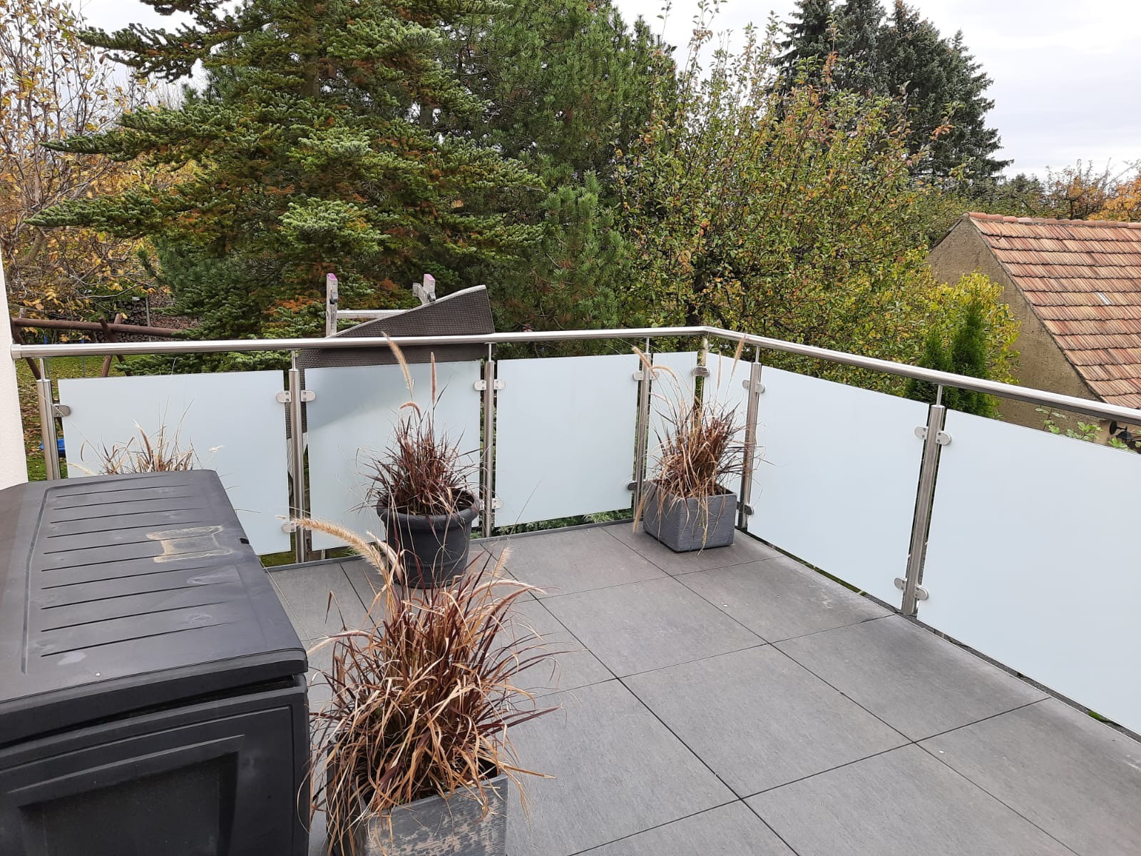 Nowoczesna balustrada balkonowa ze stali nierdzewnej i matowego szkła, z donicami z ozdobnymi trawami na szarych płytkach. Widok na zieleń drzew i fragment dachu.