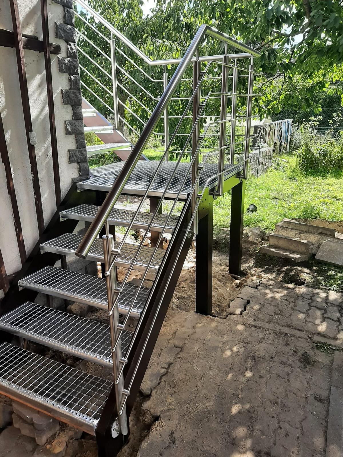 Zewnętrzne schody metalowe z ażurowymi stopniami i balustradą ze stali nierdzewnej, prowadzące do budynku z elementami kamiennymi na elewacji. Widok na ogród w tle.