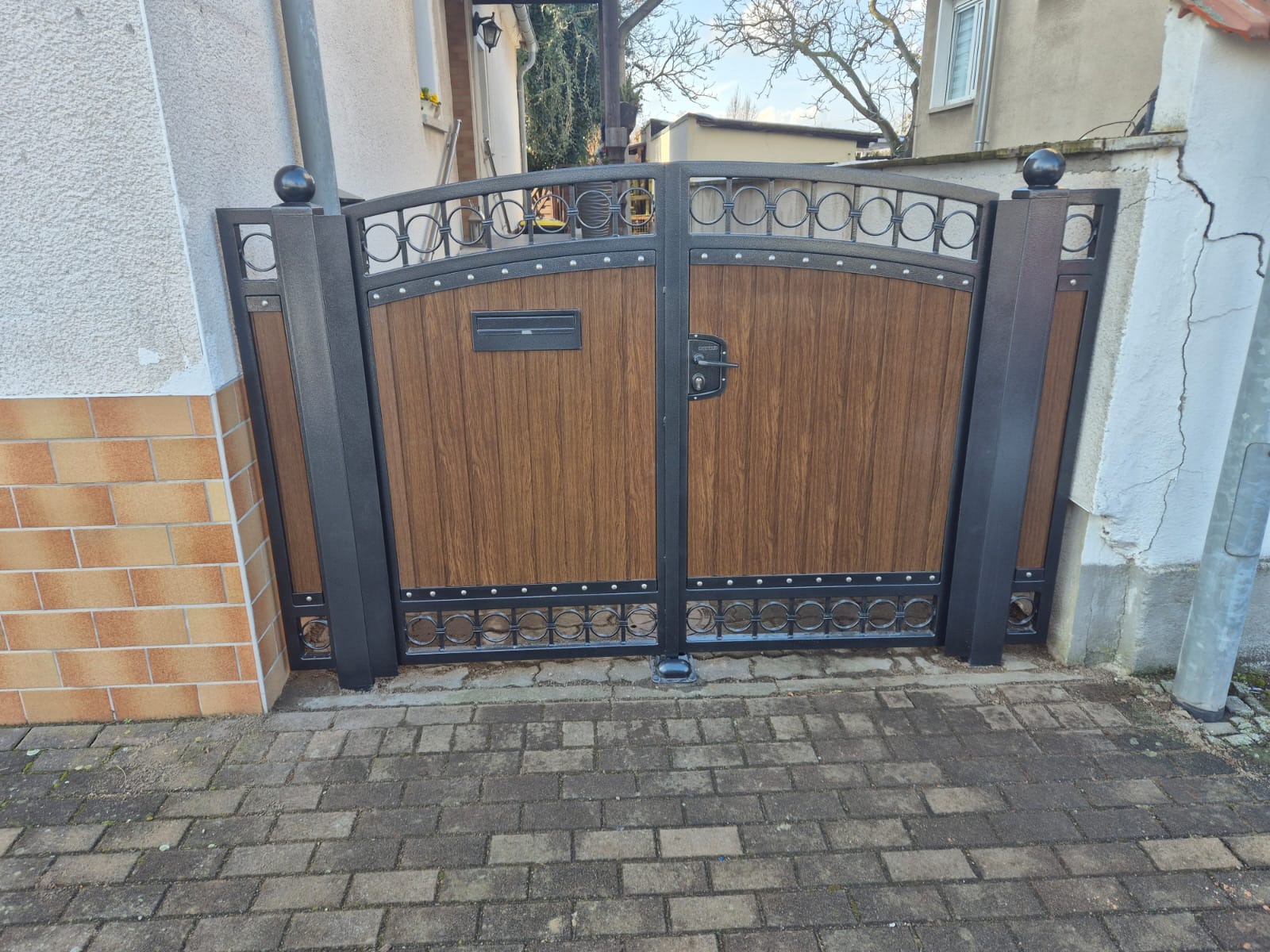 Dwuskrzydłowa brama z drewnopodobnym wypełnieniem i metalową ramą z dekoracyjnymi elementami. Skrzynka na listy wbudowana w jedno ze skrzydeł.