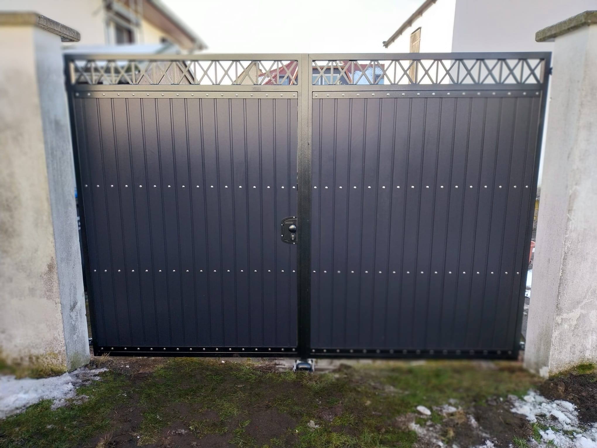 Nowoczesna, dwuskrzydłowa brama w kolorze grafitowym z dekoracyjnym, ażurowym wykończeniem u góry, zamontowana między betonowymi słupkami. Widoczne ślady śniegu.