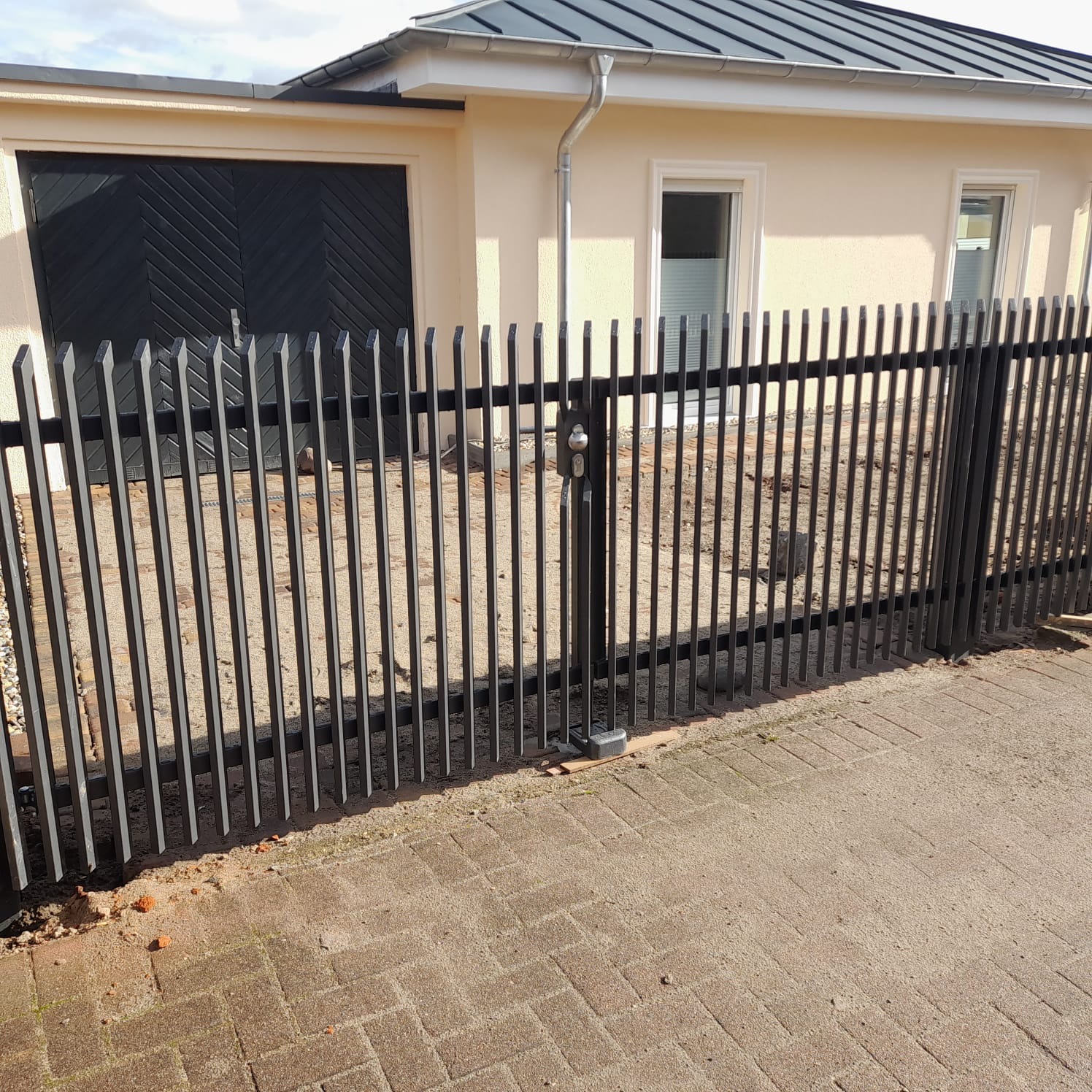 Nowoczesne, czarne ogrodzenie metalowe z pionowych prętów, z furtką zamykaną na klucz, na tle beżowego domu z czarną bramą garażową i kostki brukowej.