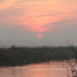 Justyna Woźna - Mglisty krajobraz z rzeką i zachodzącym słońcem, które częściowo zasłania wieża energetyczna. Delikatne, pastelowe barwy nieba odbijają się w wodzie.