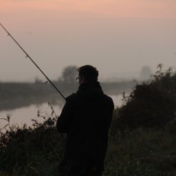 Justyna Woźna - Sylwetka wędkarza z wędką nad brzegiem rzeki o świcie, mgła nad wodą, spokojny krajobraz, stonowana kolorystyka, widok z tyłu, osoba w okularach.