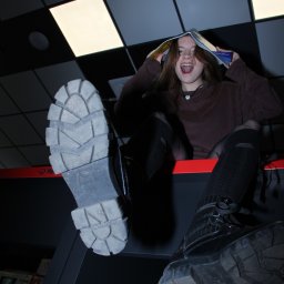 Justyna Woźna - Ekscentryczne ujęcie młodej kobiety z otwartą książką na głowie, siedzącej w pralni. Buty na pierwszym planie, nietypowa perspektywa, ekspresyjna mina.