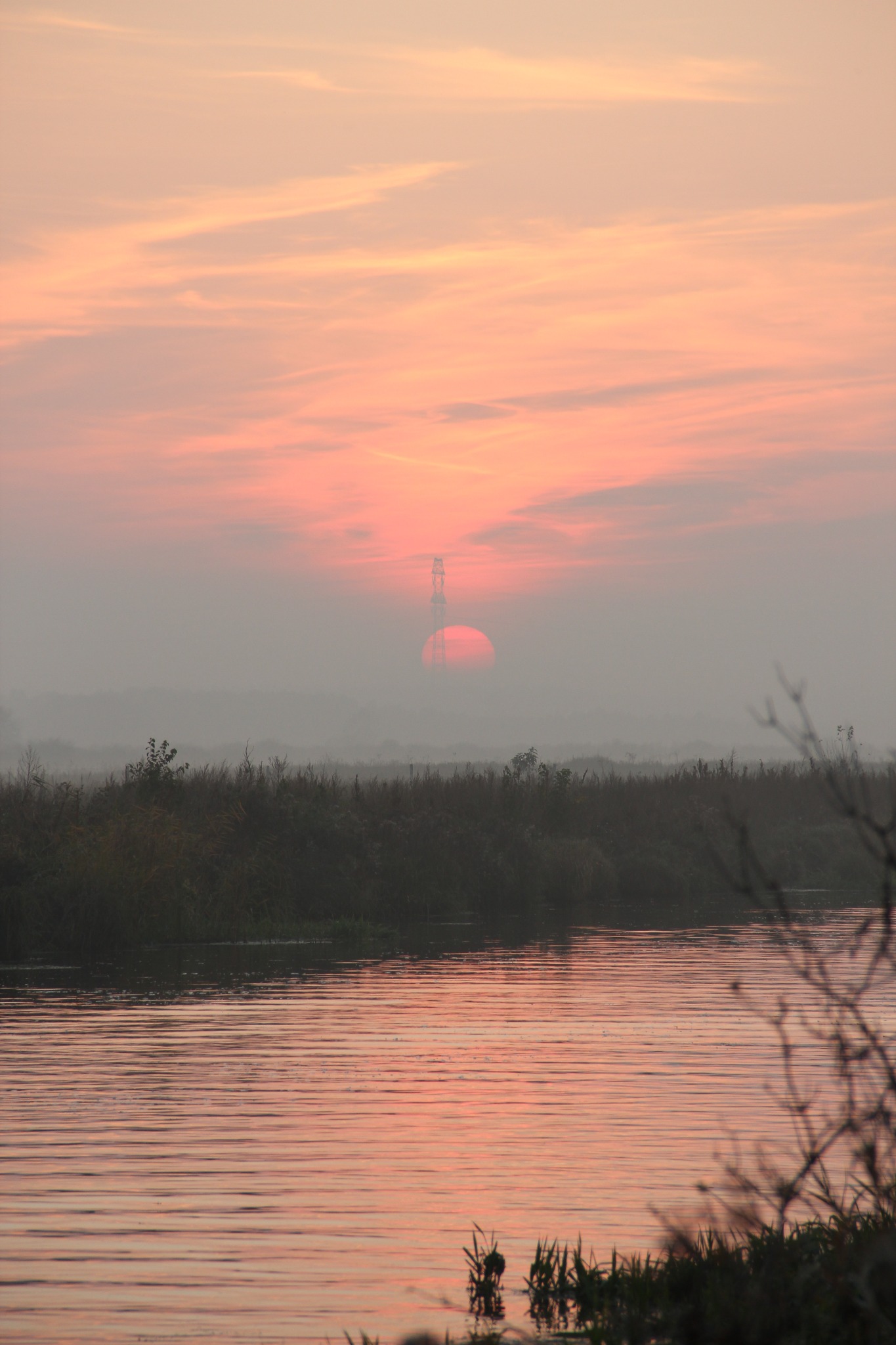 Mglisty krajobraz z rzeką i zachodzącym słońcem, które częściowo zasłania wieża energetyczna. Delikatne, pastelowe barwy nieba odbijają się w wodzie.
