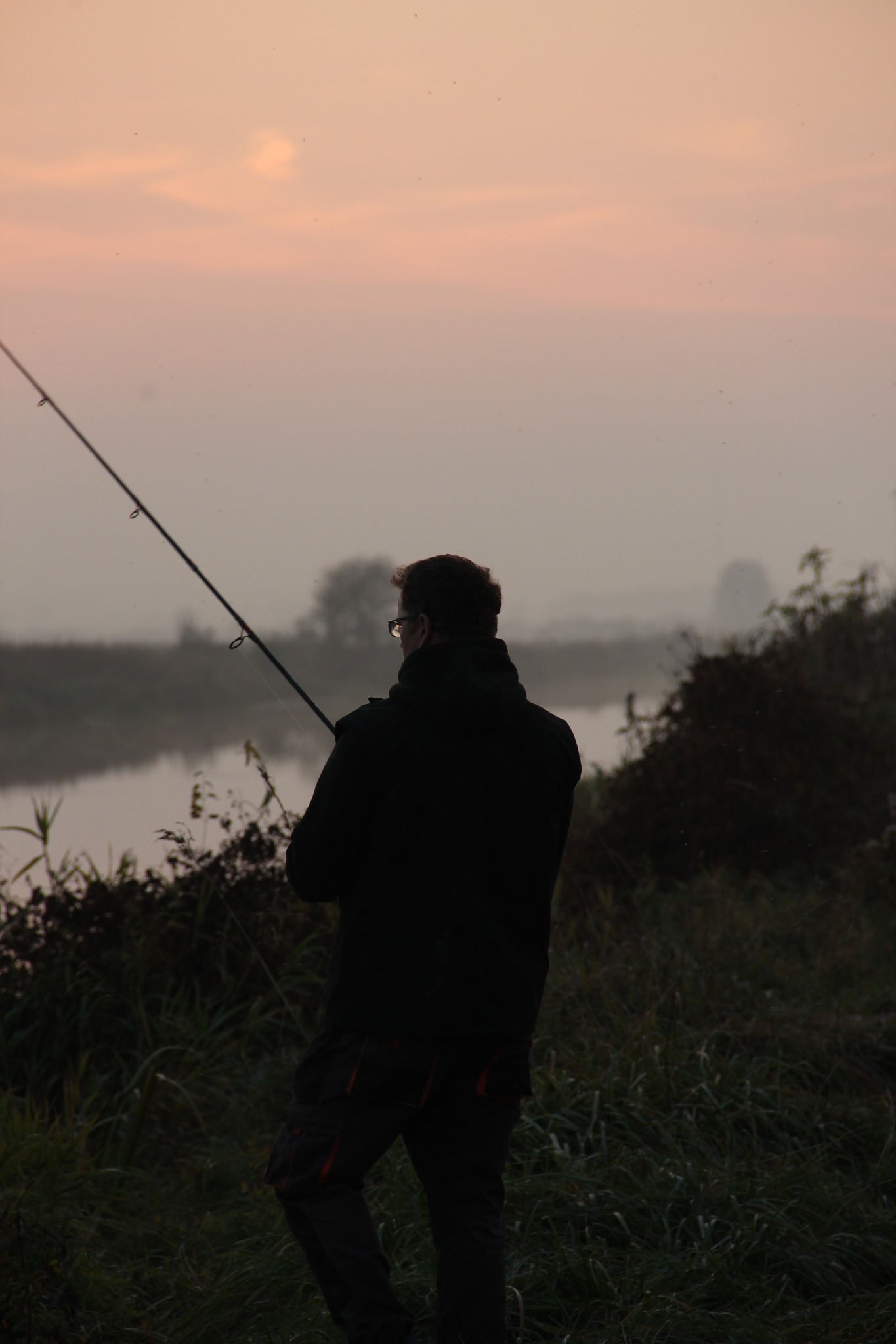 Sylwetka wędkarza z wędką nad brzegiem rzeki o świcie, mgła nad wodą, spokojny krajobraz, stonowana kolorystyka, widok z tyłu, osoba w okularach.