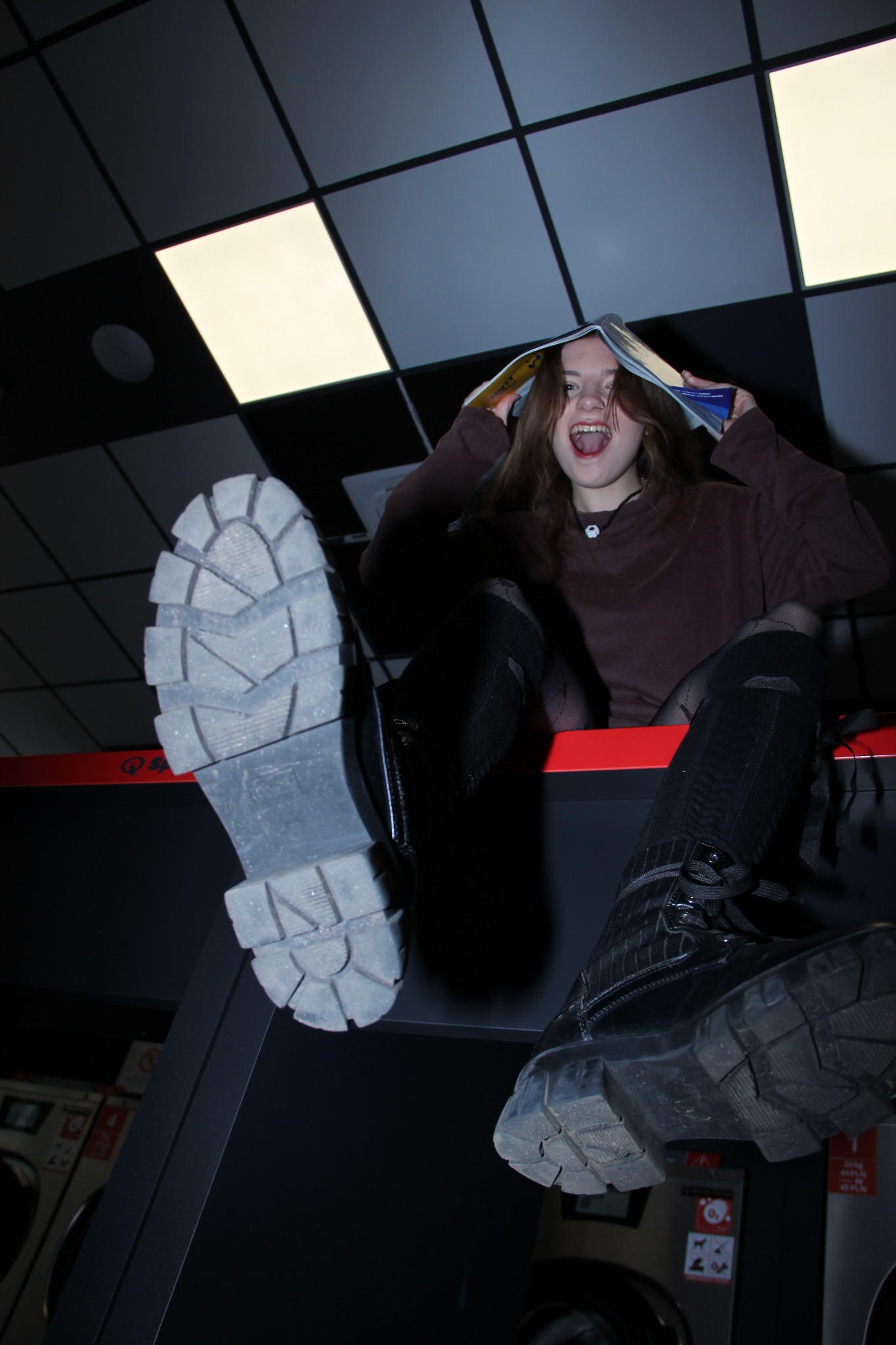 Ekscentryczne ujęcie młodej kobiety z otwartą książką na głowie, siedzącej w pralni. Buty na pierwszym planie, nietypowa perspektywa, ekspresyjna mina.