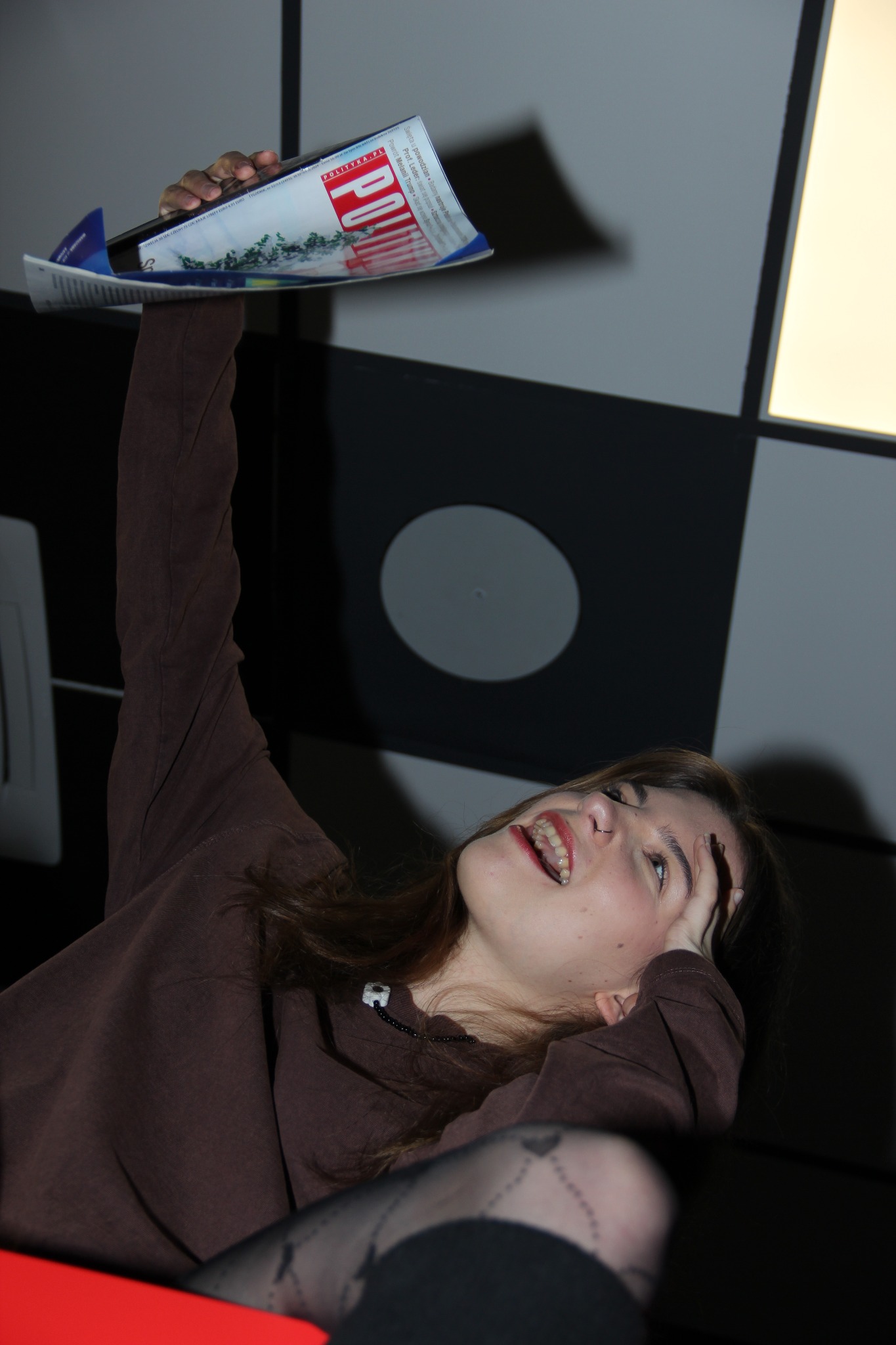 Młoda kobieta w brązowej bluzie, leżąc, trzyma nad głową otwarty magazyn 'Polityka' na tle geometrycznej ściany. Uśmiech, rajstopy w serduszka.