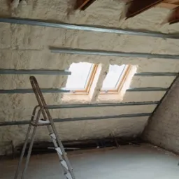 Poddasze z oknami dachowymi w trakcie ocieplania pianą poliuretanową, widoczne metalowe profile montażowe oraz drabina malarska.