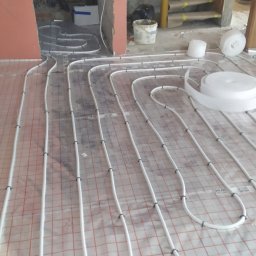 Artur Podgórski BUDOTUR - Rozłożona instalacja ogrzewania podłogowego na folii z siatką, widoczne rury mocowane spinkami, fragment ściany w stanie surowym oraz rolka taśmy montażowej.