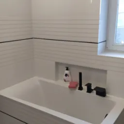 Nowoczesna, biała wanna z czarną armaturą i półką z kosmetykami w łazience wykończonej białymi płytkami z poziomymi liniami.