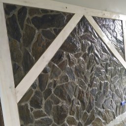 Artur Podgórski BUDOTUR - Ściana wyłożona nieregularnym kamieniem w odcieniach szarości i brązu, z drewnianą ramą w jasnym odcieniu, tworzącą geometryczny wzór na powierzchni