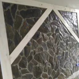 Ściana wyłożona nieregularnym kamieniem w odcieniach szarości i brązu, z drewnianą ramą w jasnym odcieniu, tworzącą geometryczny wzór na powierzchni