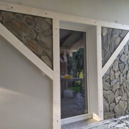 Artur Podgórski BUDOTUR - Fragment elewacji budynku z wykończeniem z kamienia naturalnego w drewnianej ramie, widoczny przez okno stół z żółtą miską i wiadro, na podłożu folia ochronna.