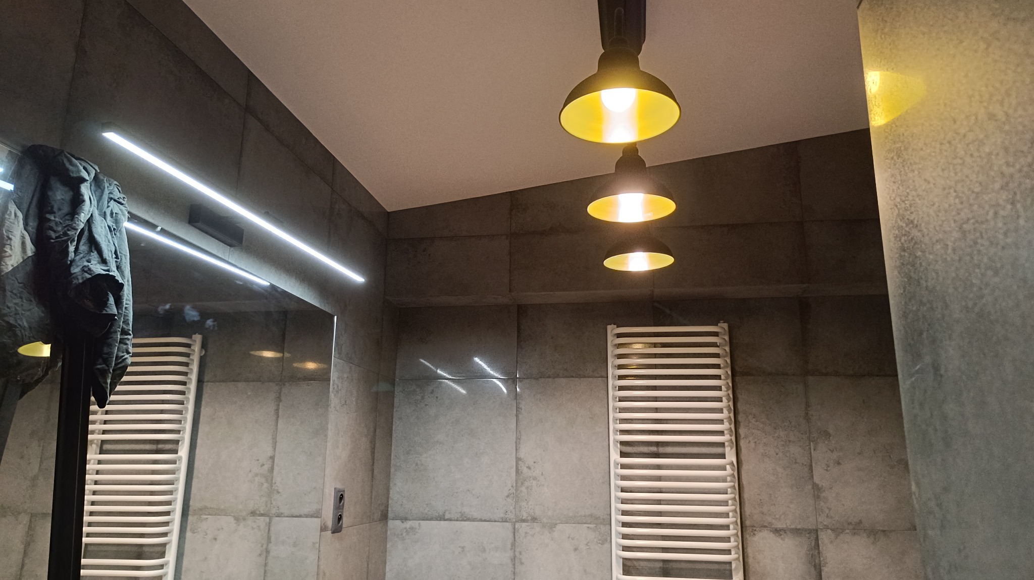 Szara łazienka z trzema lampami w stylu industrialnym, grzejnikiem drabinkowym i podświetlanym lustrem, na którym wisi kurtka.