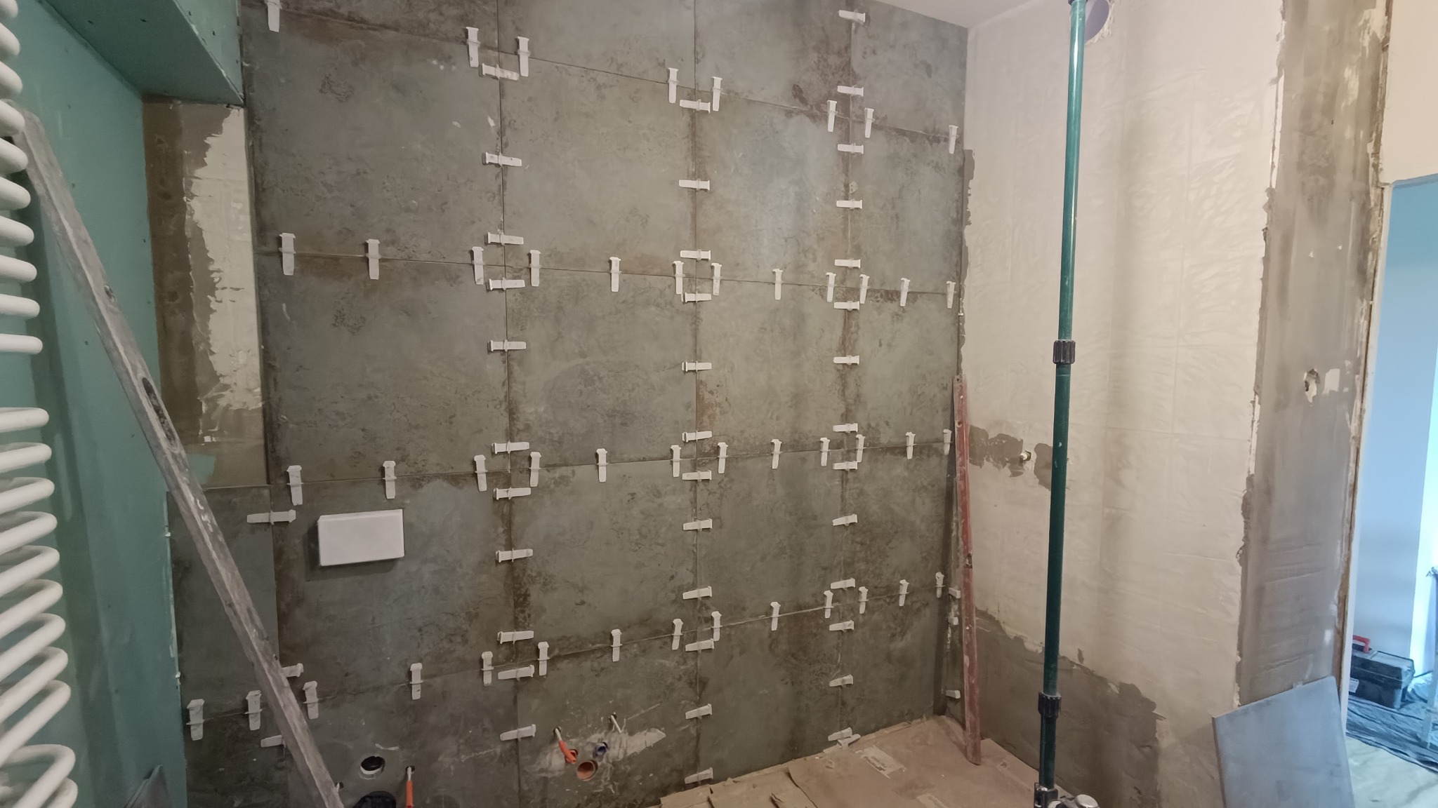 Ściana w trakcie wykańczania z szarymi płytkami, widoczne krzyżyki dystansowe, fragment grzejnika drabinkowego i poziomica, ściana częściowo otynkowana.