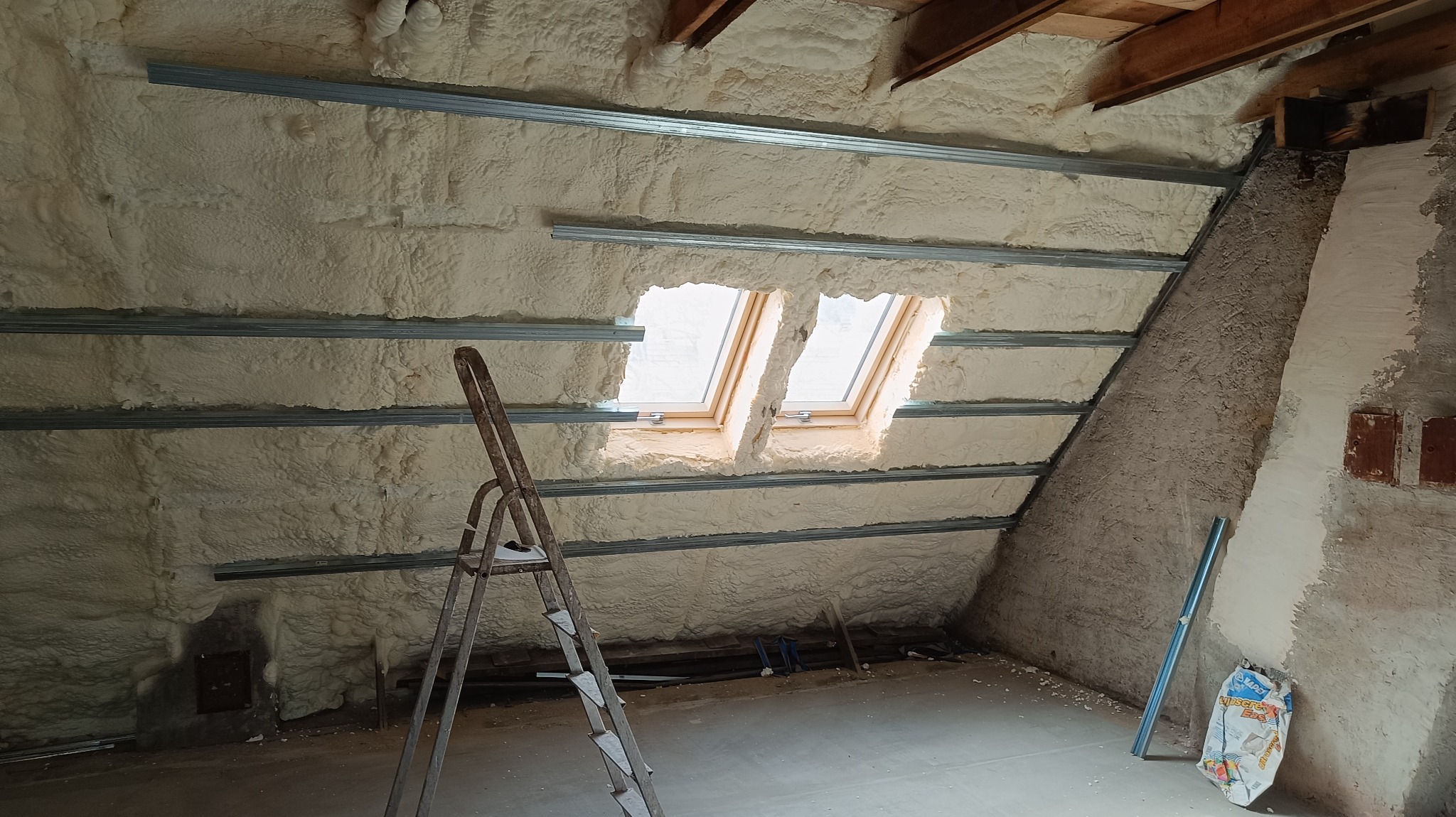Poddasze z oknami dachowymi w trakcie ocieplania pianą poliuretanową, widoczne metalowe profile montażowe oraz drabina malarska.