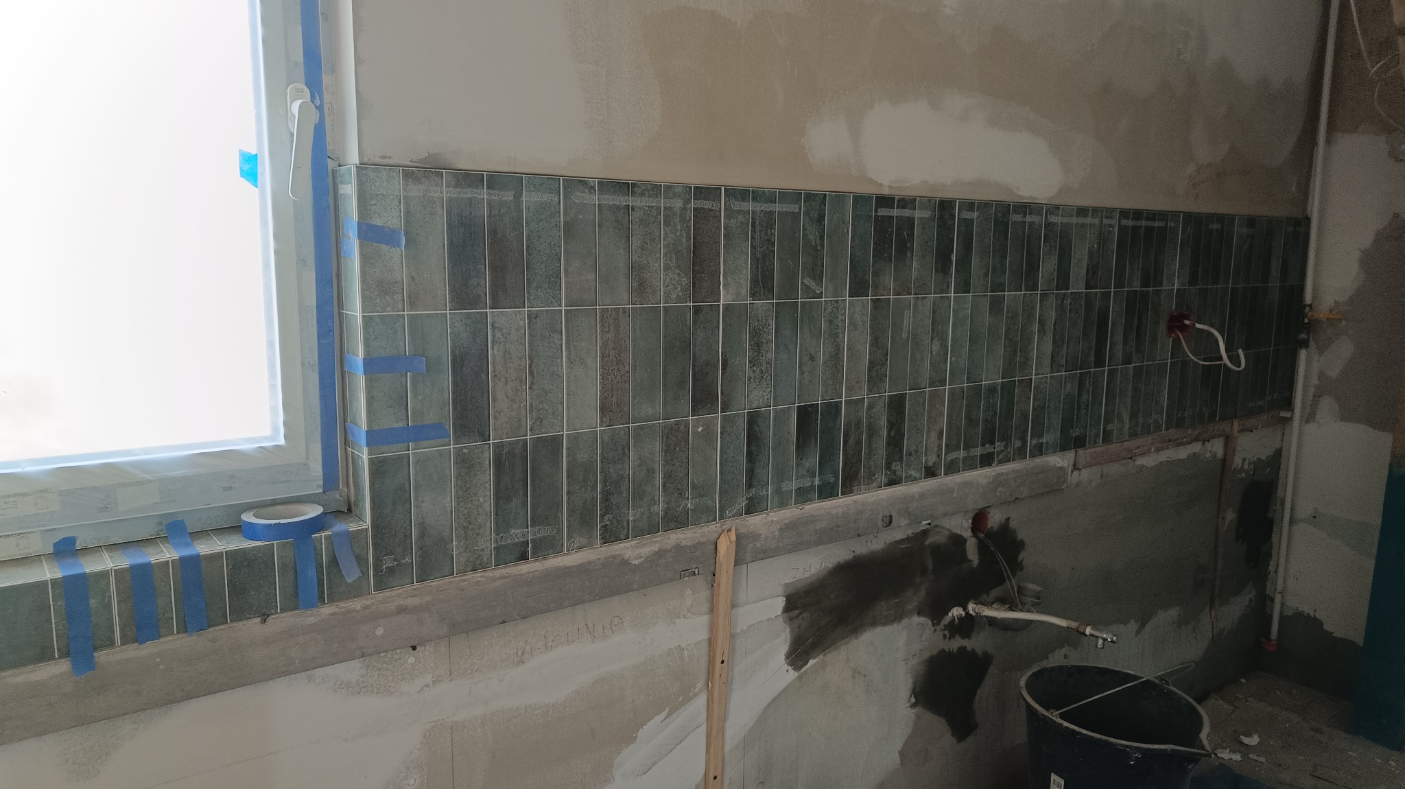 Układanie pionowych, smukłych, zielono-szarych płytek na ścianie w trakcie prac wykończeniowych, z widocznym oknem zabezpieczonym folią i niebieską taśmą malarską oraz elementami instalacji...