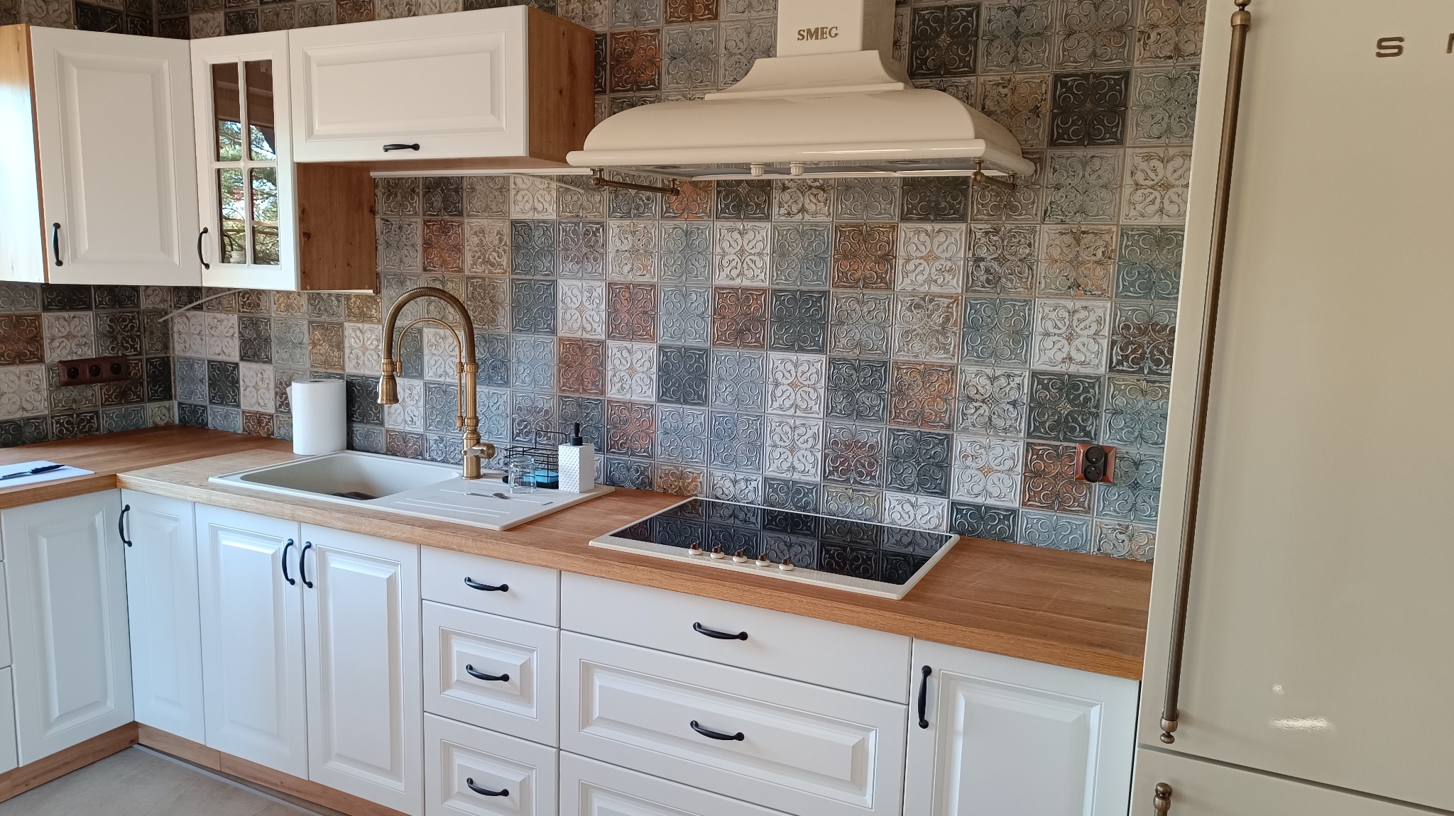 Nowoczesna kuchnia z białymi szafkami, drewnianym blatem, ozdobnymi płytkami na ścianie i złotą baterią przy zlewie.