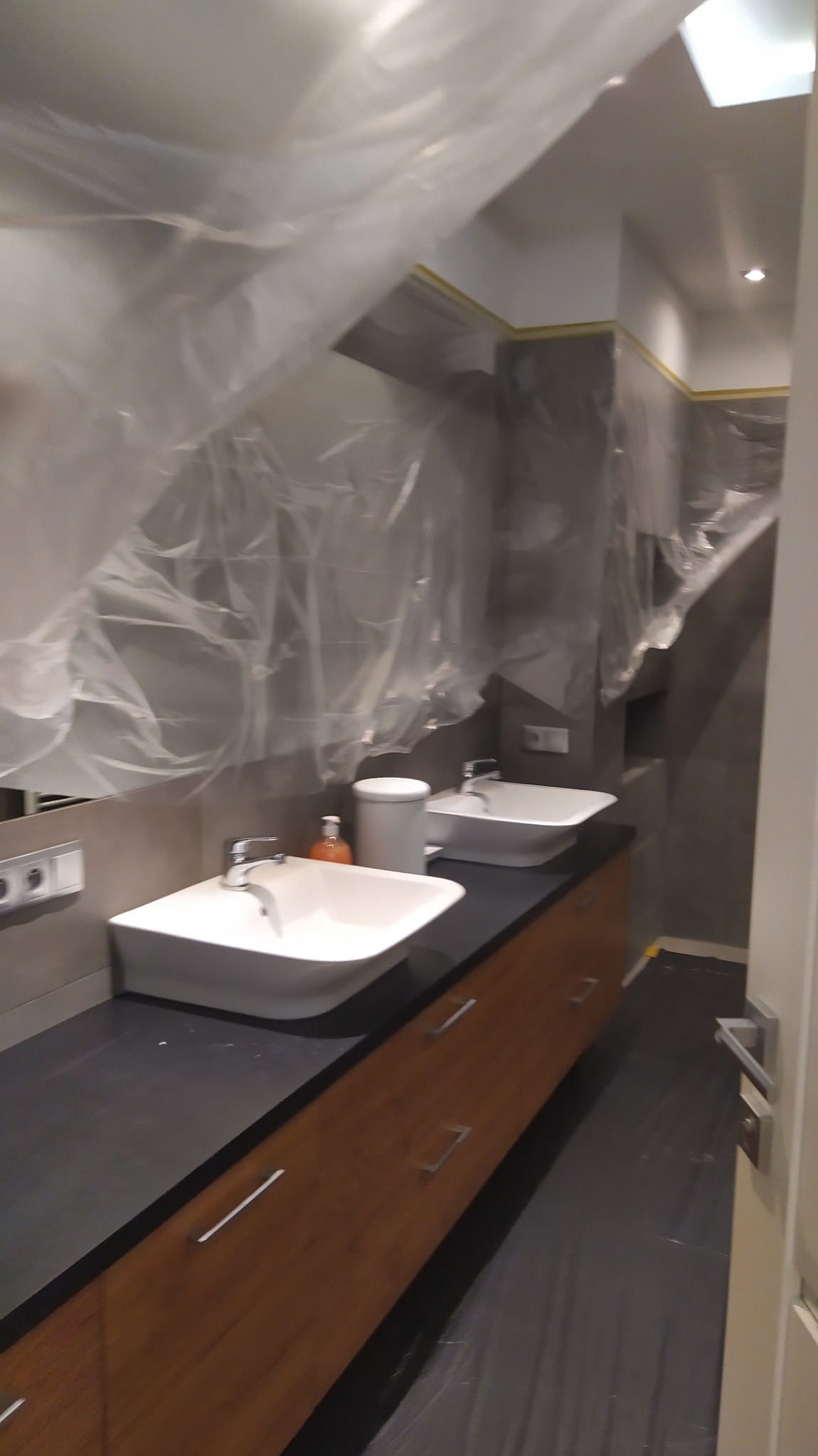Łazienka w trakcie prac wykończeniowych, zabezpieczona folią malarską, widoczne dwie umywalki nablatowe na szafce z drewnopodobnym frontem i czarnym blatem, szare płytki na ścianach.